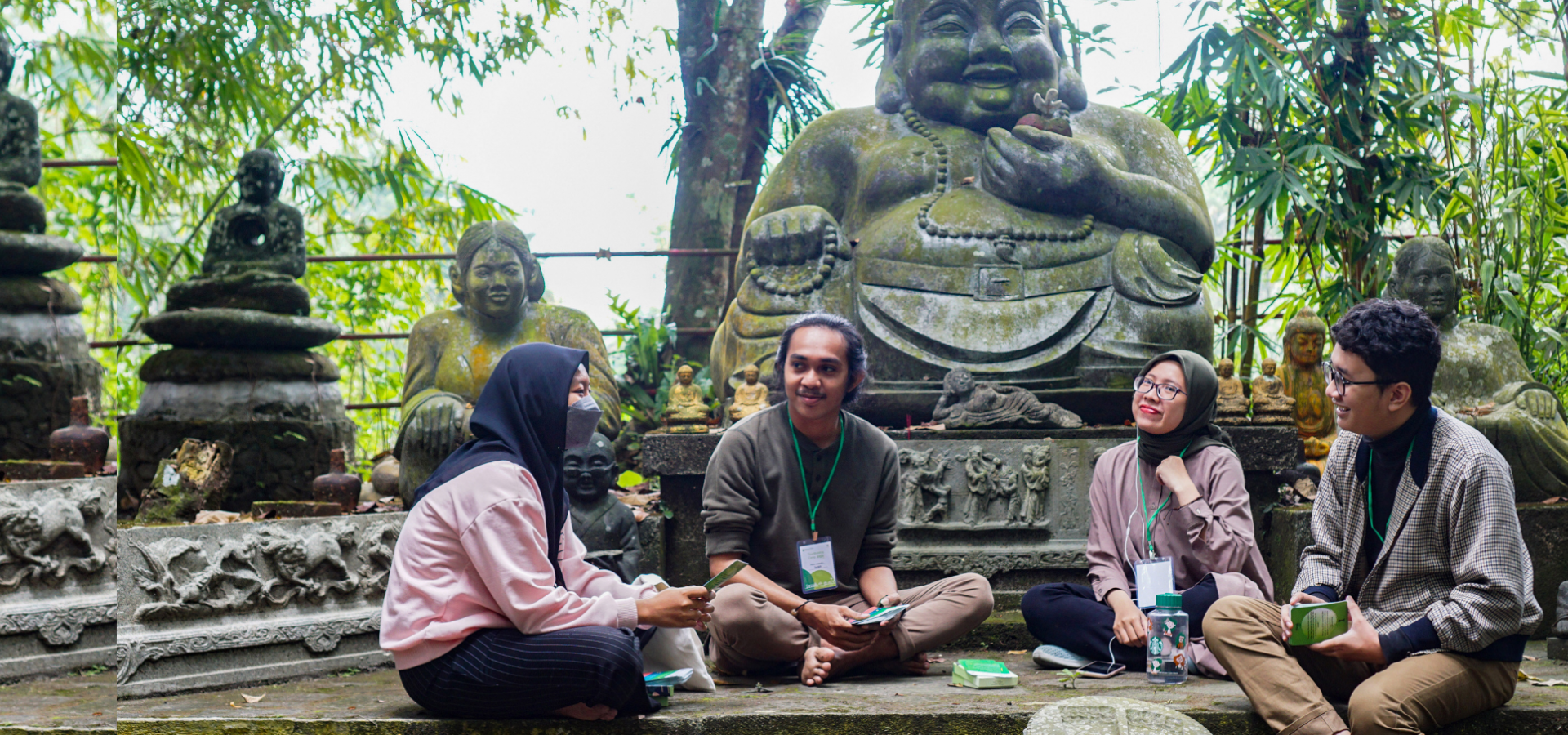 Four Indonesian youth sit in front of a stone statue surrounded by lush greenery, talking