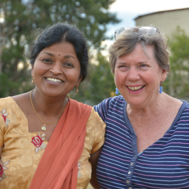South Asian woman in sari and white woman in blue tee-shirt stand shoulder to shoulder and smile at the camera