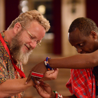 White male with glasses and blond beard receives a bracelet from a Black man wearing African shawl