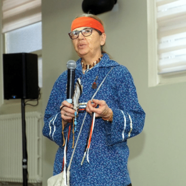 Indigenous Canadian woman, wearing blue tunic and orange headband, speaks before a crowd