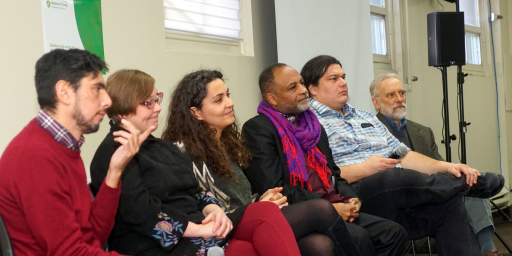Six panelists, of different ages, genders, and ethnicities, look out over crowd of attendees,