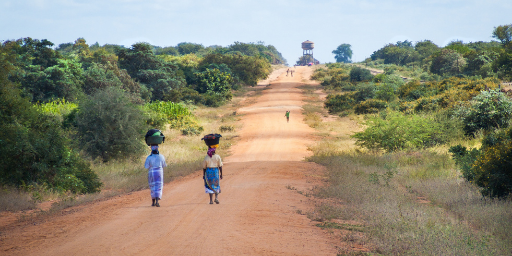 Two African women with baskets on their heads walk down a long, dusty road