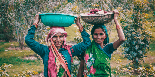 Two southeast asian women, balancing bowls on their heads, look out over a field and smile at one another